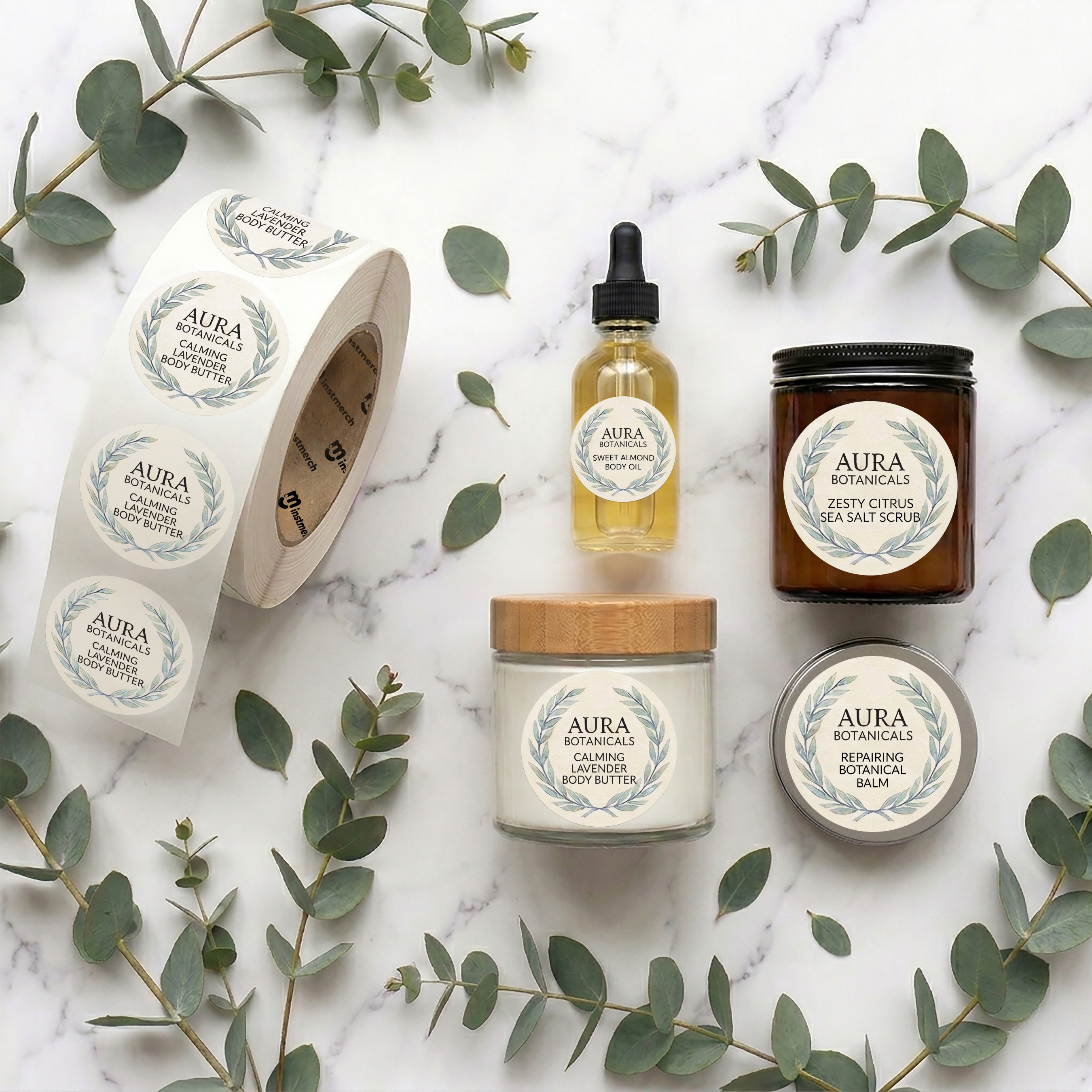 A flat-lay arrangement of AURA Botanicals natural body care products on a white marble background with eucalyptus leaves. The products include a roll of stickers, a dropper bottle of essential oil, a jar of sea salt scrub, a candle, and a tin of body butter.